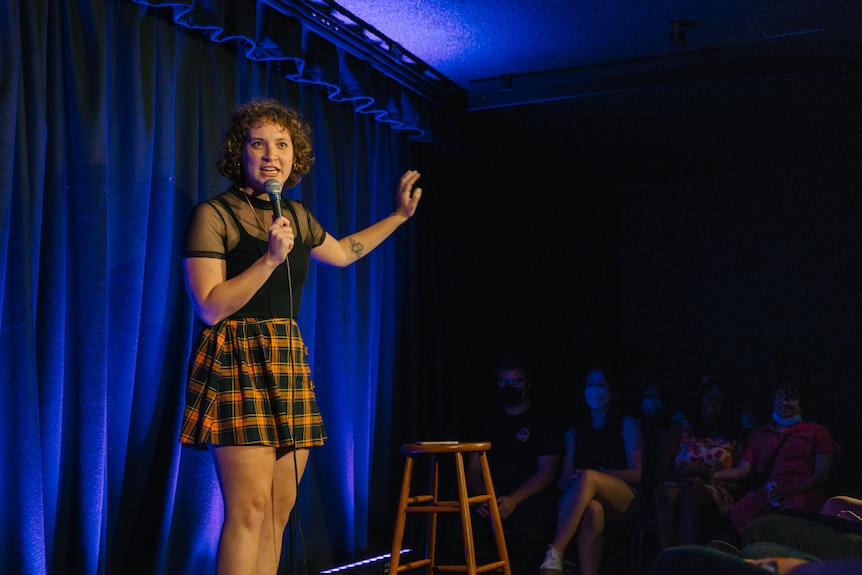 A comedian holds a mic on stage in front of a stool, gesturing for emphasis