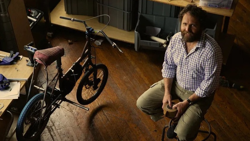 A bearded middle-aged man sits on a stool beside a bike