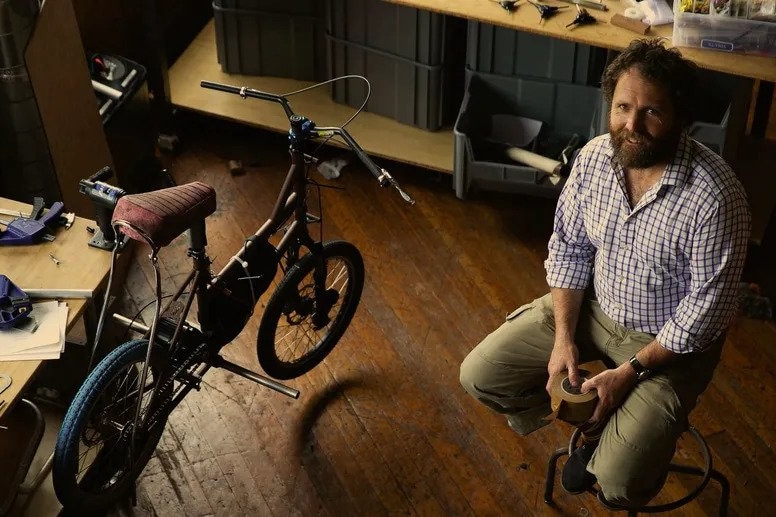 A bearded man sits on a stool beside a bike