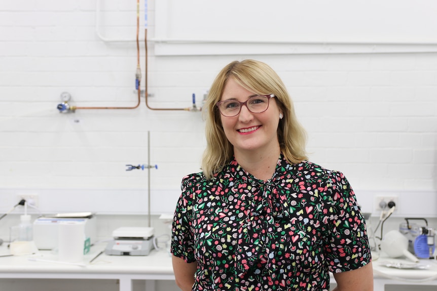 Smiling Caucasian woman, blonde, wears multicoloured black top, glasses, in a lab, white wall behind.