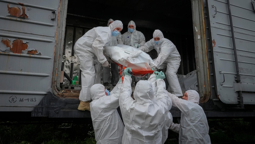 Ukrainian servicemen load bodies of Russian soldiers in to a railway refrigerator carriage. They are wearing white hazmat suits.