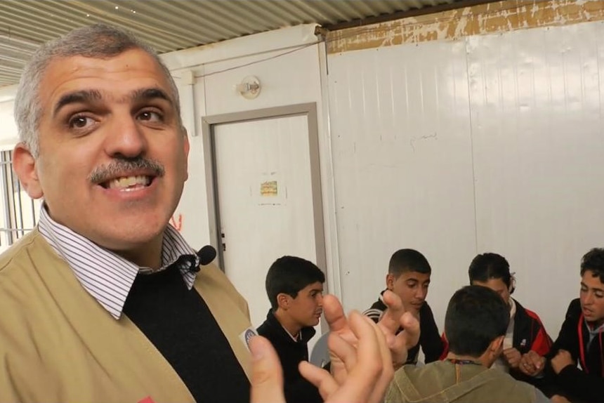 A man looks at the camera as children sit in a circle in the background.