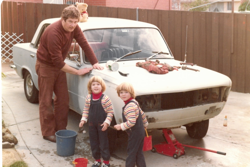 Man stands with hand on car, toddler twins stand in foreground in matching overalls and stripey tops