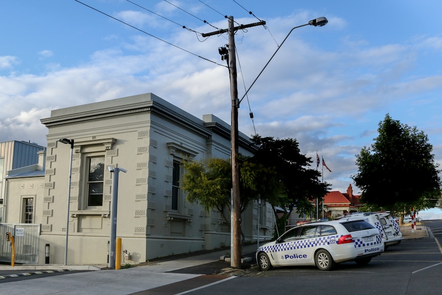 Street view of police cells, with police cars parked out front on tree-lined street.