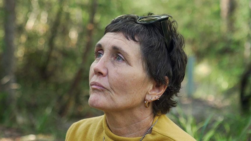 A woman with short dark hair is looking off into the distance in front of a forest