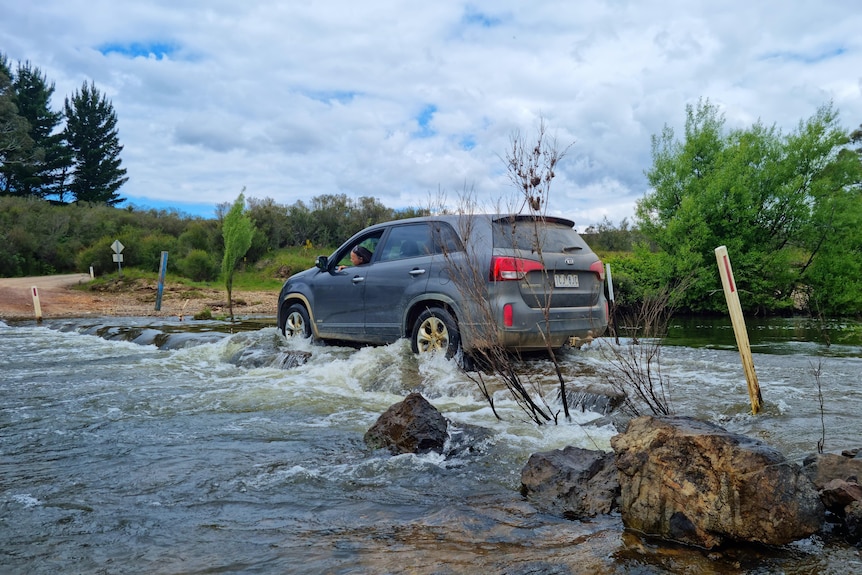 A car drives across a rushing causeway, covered in river water.