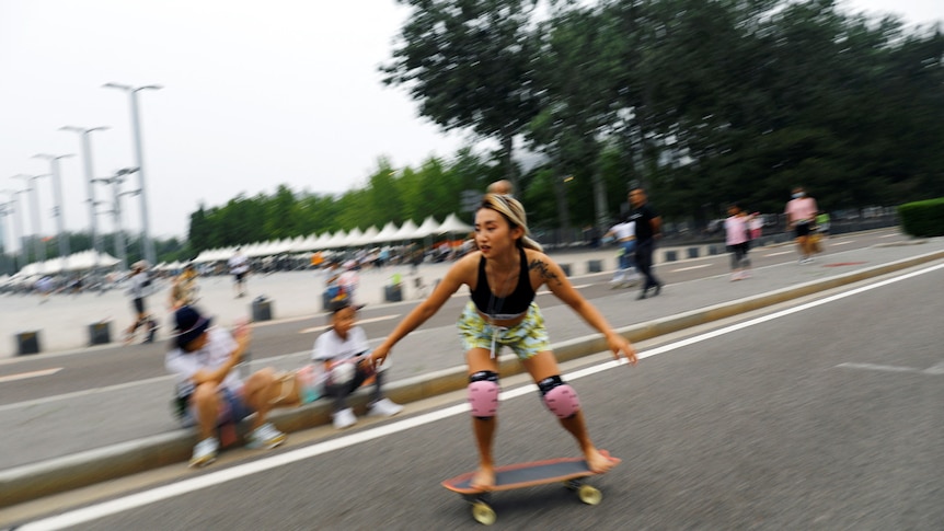 A woman rides a skateboard down a street and people watch while sitting on the curb. 