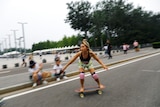 A woman rides a skateboard down a street and people watch while sitting on the curb. 