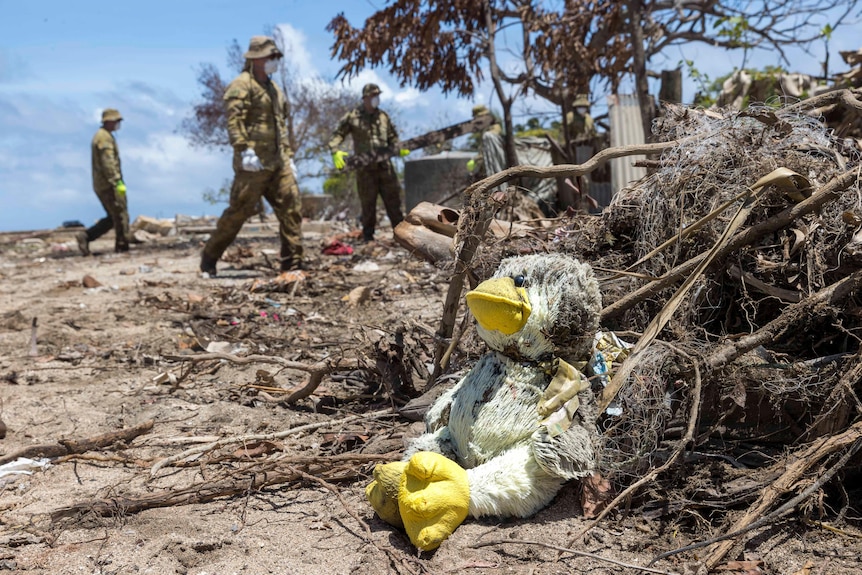 A dirty soft toy is in the foreground with soldiers walking among debris in the background.