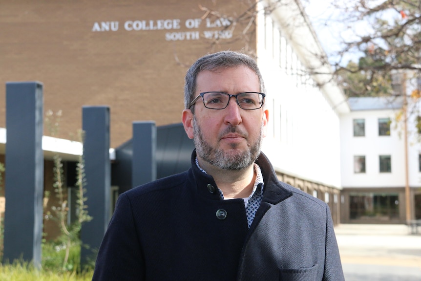 A man stares part the camera with a serious expression at the front of a university building. 