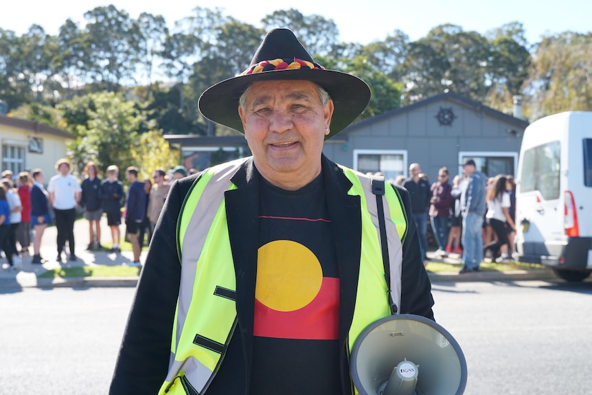 An Indigenous elder in a shirt with the Aboriginal flag on it, holding a megaphone and smiling at the camera