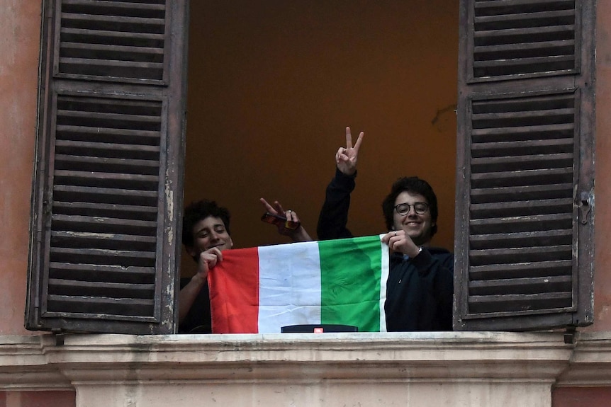 Two young Italians giving peace signs and holding an Italian flag out a window