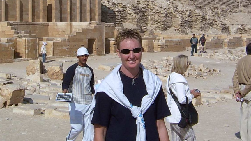 Woman wearing glasses and white jumper over black t-shirt standing in front of Egyptian pyramid.