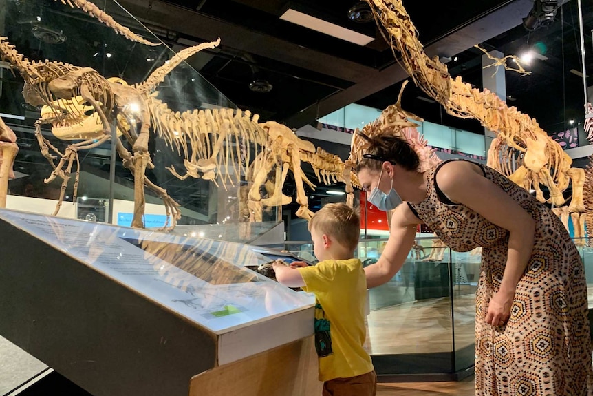 A boy and a woman wearing a face mask look at a display or dinosaur skeletons.