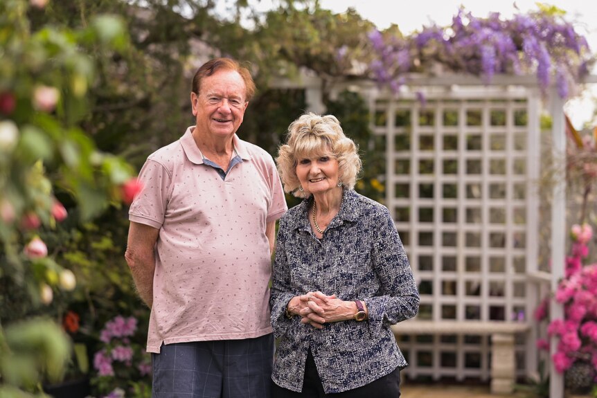 Elderly couple, a man and woman, standing next to each other in a garden smiling. 