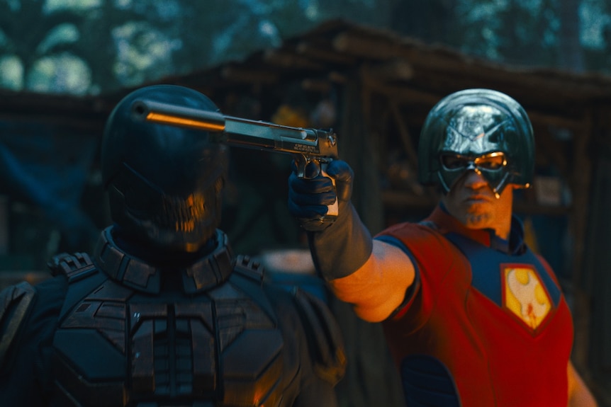 A man in a red superhero outfit, including a metal helmet, holds out a gun. Next him is another man in a black full body suit.