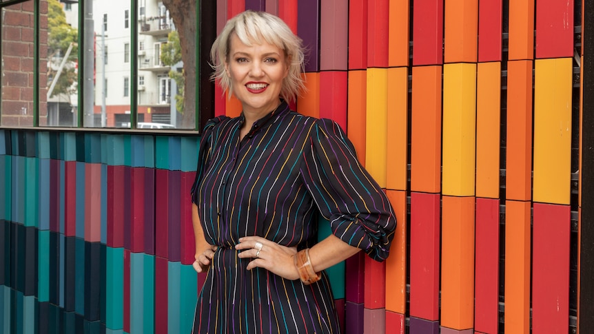 A smiling blonde woman stands with her hands on her hips in front of a colourful, modern-looking office wall.