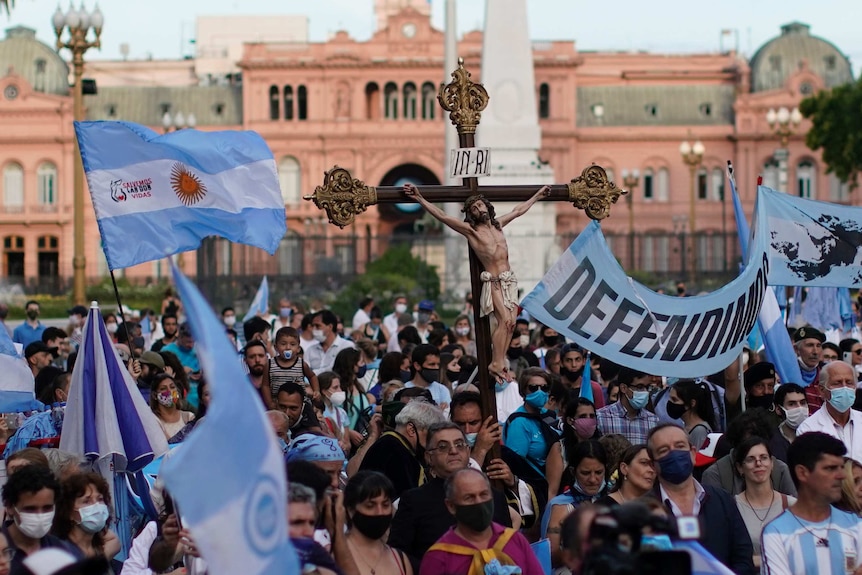 A crowd of people holding blue signs and a figure of Jesus Christ on the cross gather outside a building.