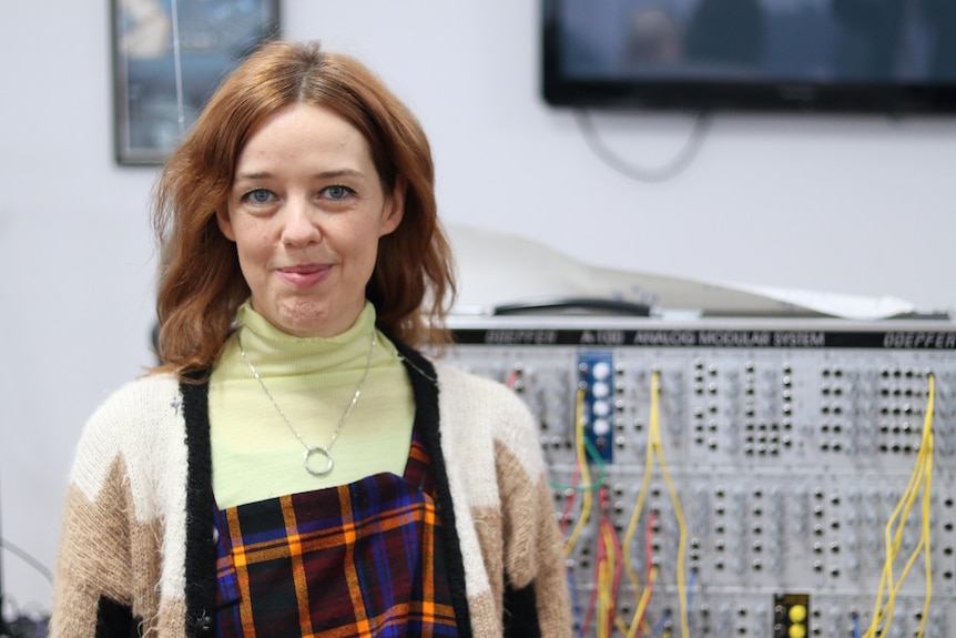 A smiling woman with shoulder-length brown hair stands in front of a computer switchboard wearing a cream cardigan.