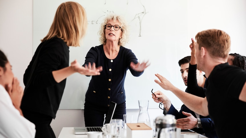 A woman tries to calm her angry colleagues in a busy boardroom
