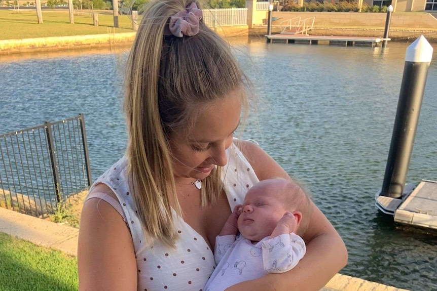 A woman holds a newborn baby, standing in front of a canal of water .