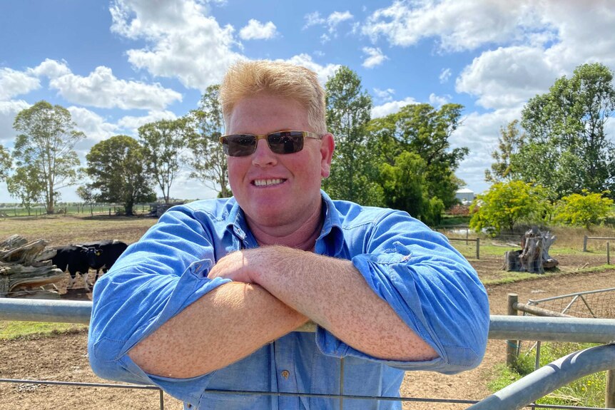 A blonde-haired man leaning on a farm gate in a paddock on a dairy farm