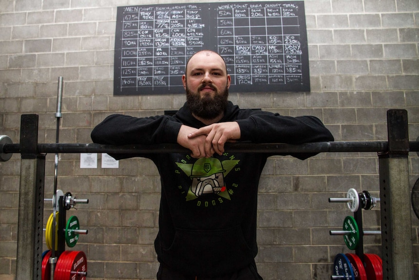 A bearded man poses at a set of barbells inside an industrial-style gym.