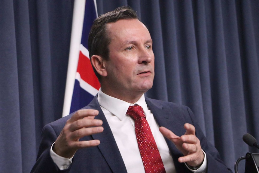 Mark McGowan addresses the media in a blue suit and red tie with the WA flag behind him