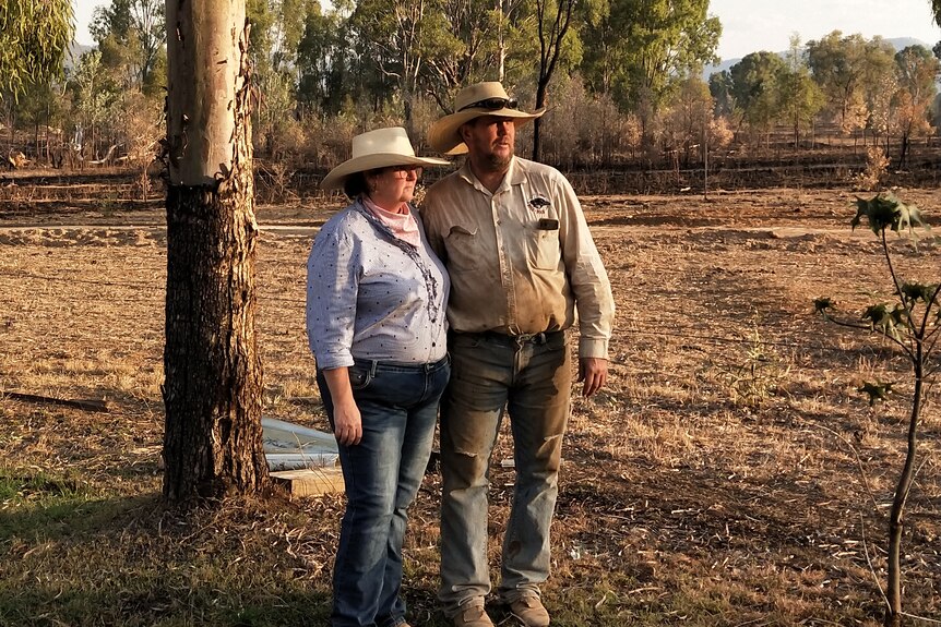 A man and a woman, both wearing akubras, shirt, jeans man covered in dust, in a burned paddock, trees in the background.