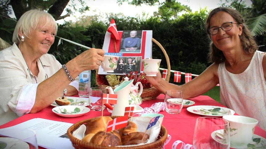 Two women bump tea cup together on a festival table in a park covered in Danish flags and food.