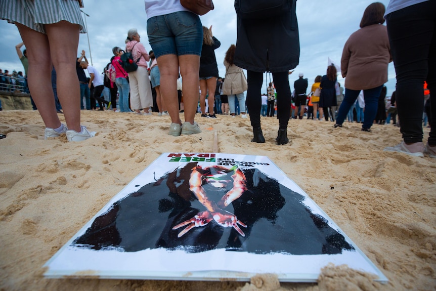 A sign rests on the sands of Bondi Beach as faceless people stand around. 