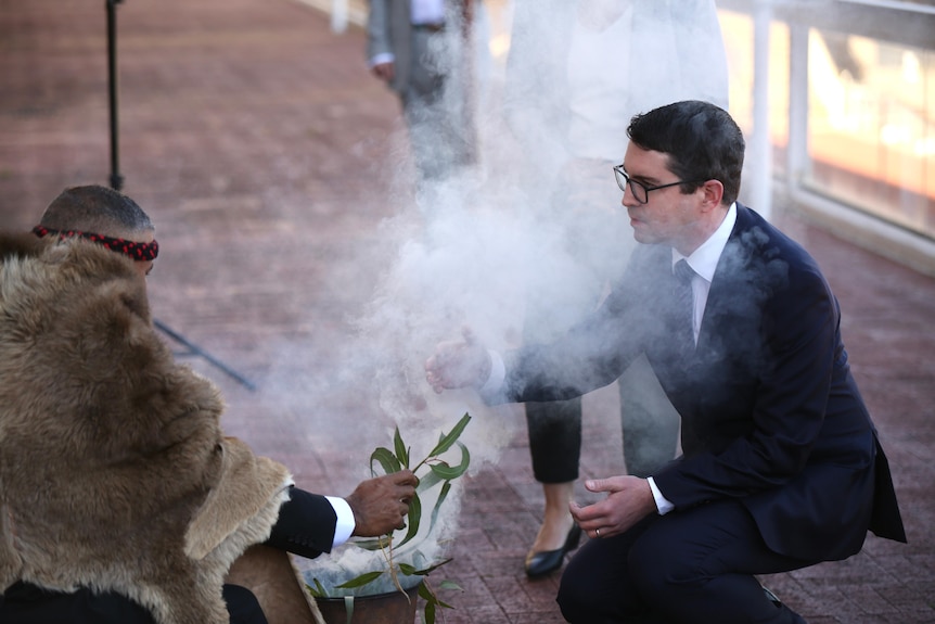 A man dressed in a suit bends down to be covered in smoke produced by an Aboriginal man. 