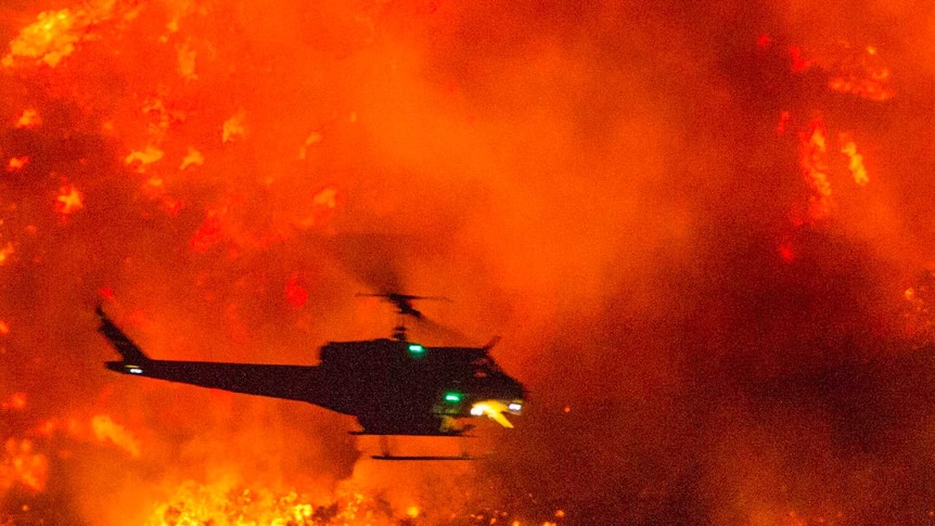 A small dark helicopter dropping water on a large backdrop of fire