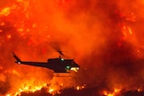 A small dark helicopter dropping water on a large backdrop of fire