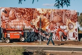 A man walks past an artist painting a large mural of flowers and seeds on a library wall