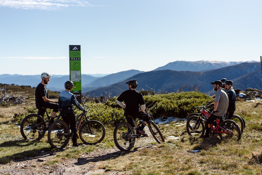 A group of people stand on their mountain bikes at the top of a mountain talking 