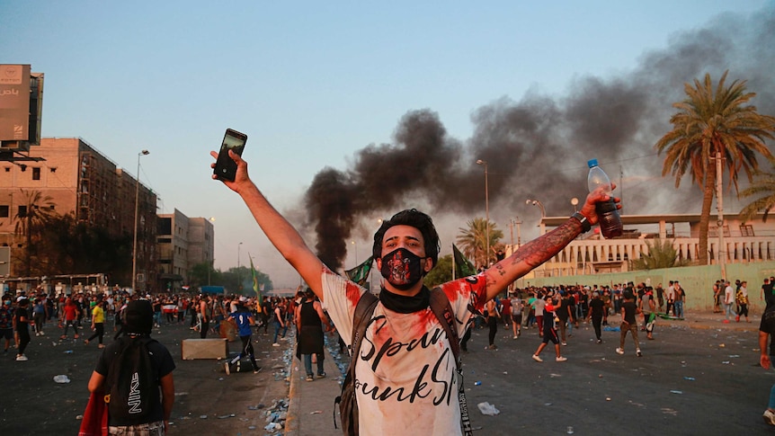 A protester with a white, blood-stained shirt stands in front of a huge crowd with his arms open in defiance at sunset.