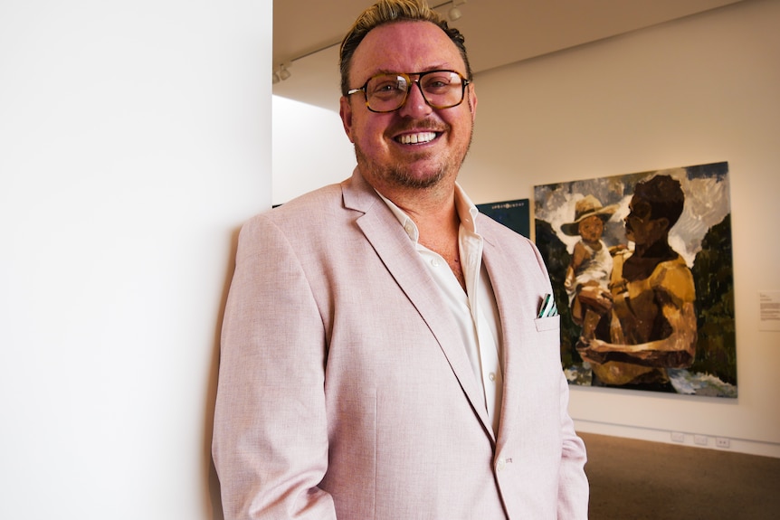 a man stands in a gallery smiling at the camera with art in the background