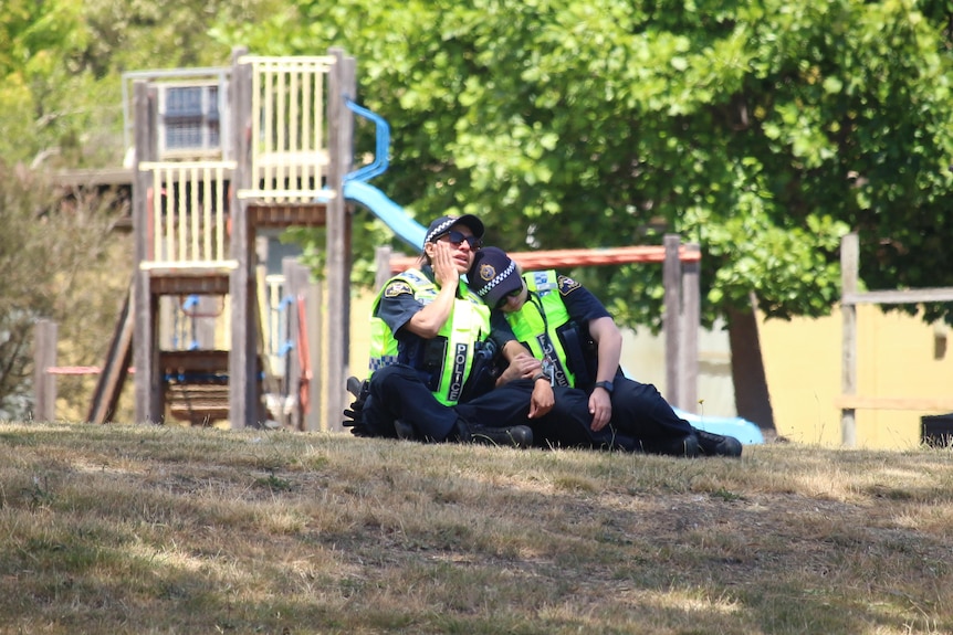 Two police officers sitting on the ground are leaning against each other.