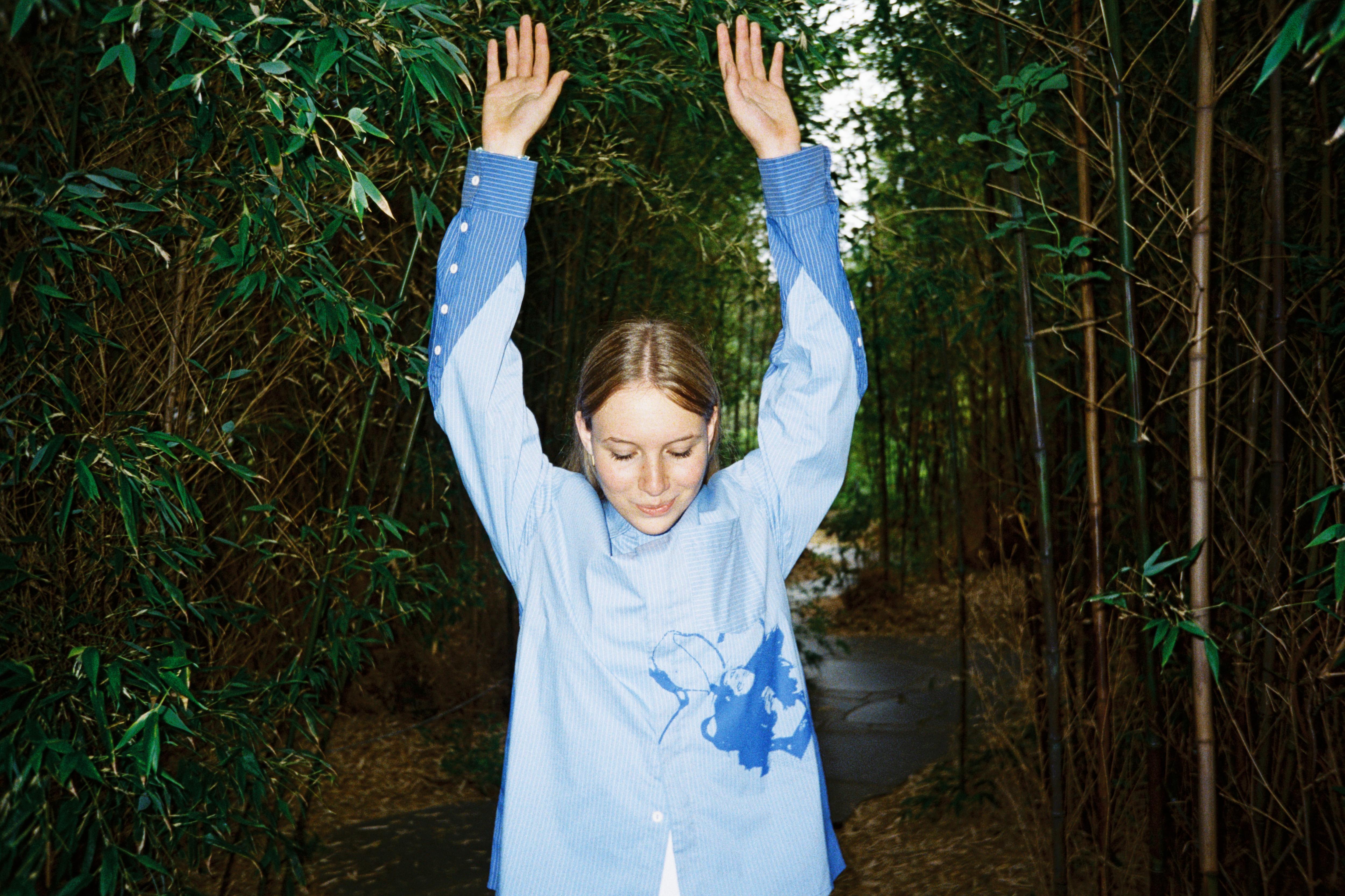 A woman in a blue dress blouse with her hair pulled back looks down with her arms above her head.