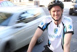 Upper body photo of Mark Bailey, wearing bike helmet and lycra, stopped beside a road as a car, blurred, drives past.