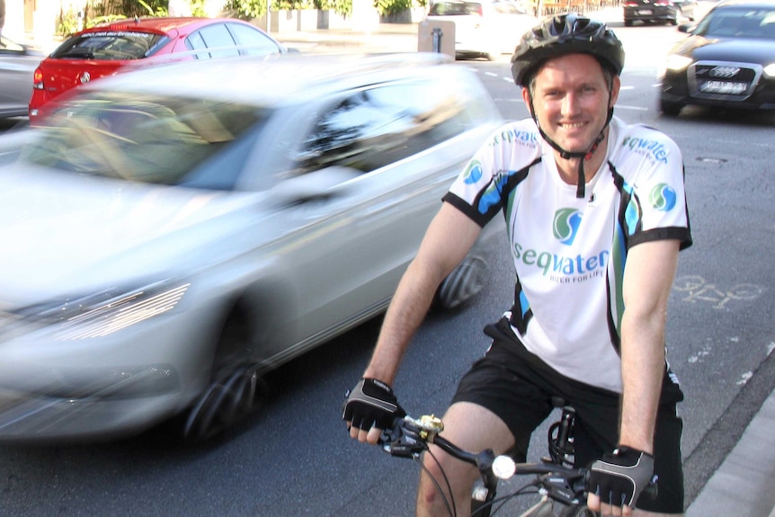 Upper body photo of Mark Bailey, wearing bike helmet and lycra, stopped beside a road as a car, blurred, drives past.