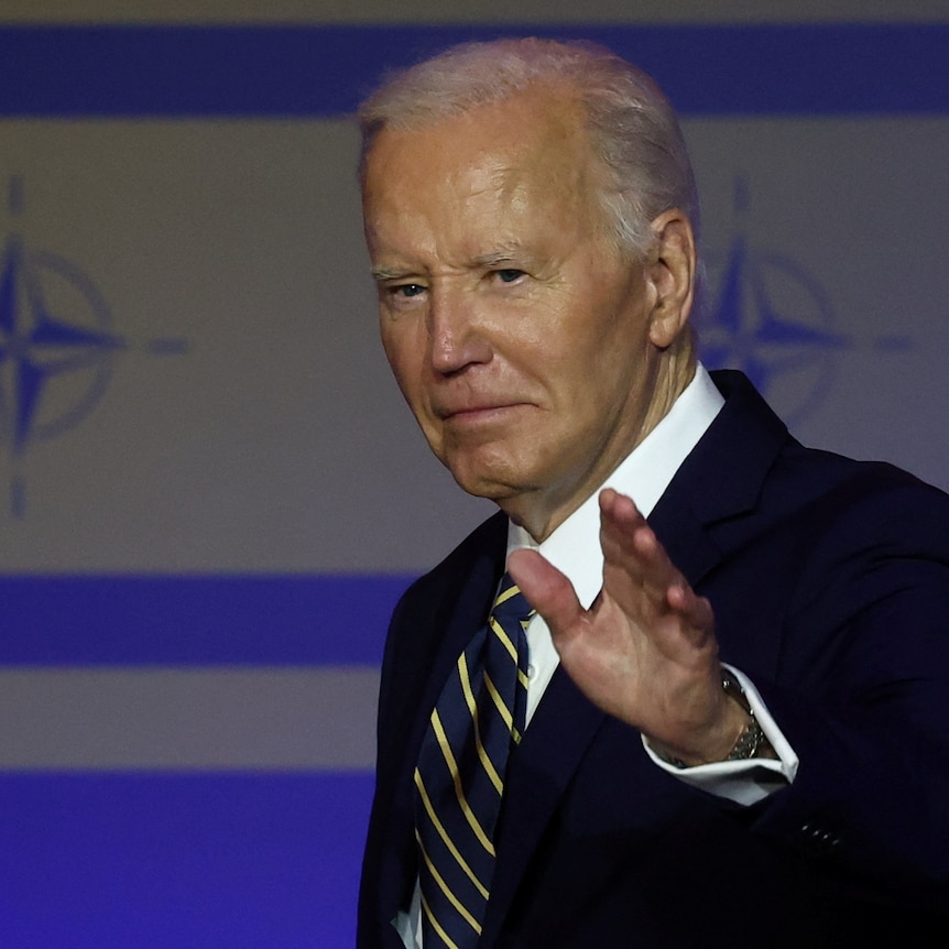 Joe Biden waves at the camera, looking serious, the NATO logo is imprinted on the wall behind him
