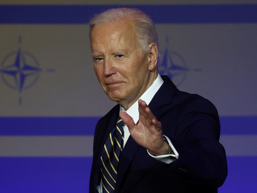 Joe Biden waves at the camera, looking serious, the NATO logo is imprinted on the wall behind him