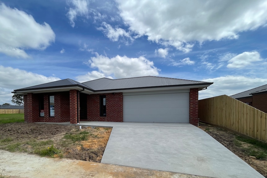 A red brick home at the top of a driveway. There is a garden bed to the left which is unfinished. 