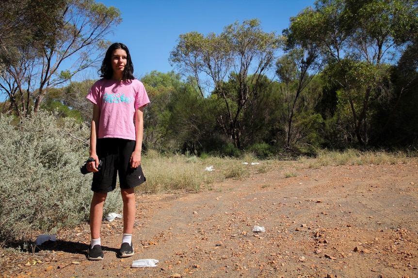 A teenage girl with dark hair standing in bushland surrounded by discarded rubbish.