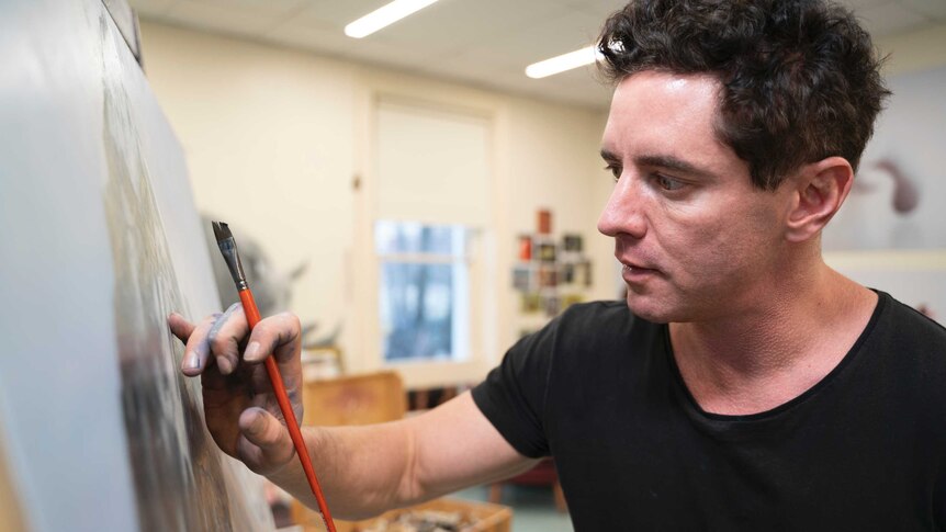 A man in a black tshirt holds a paintbrush while working on a painting