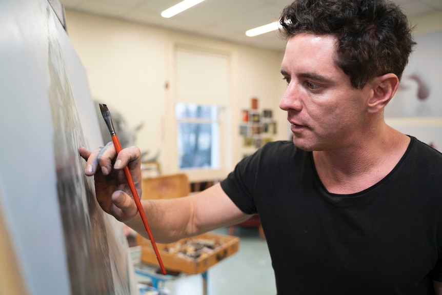 A man in a black tshirt holds a paintbrush while working on a painting