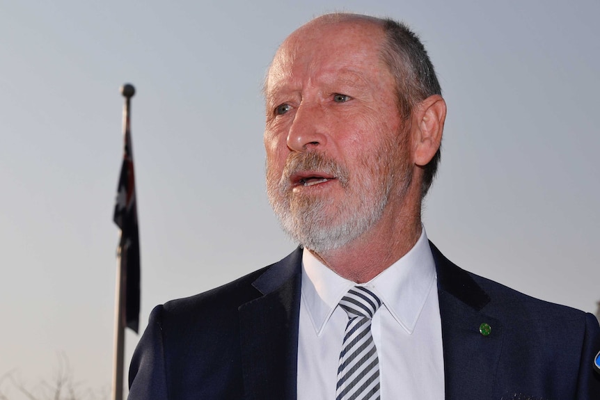 A man in a dark suit with a light grey beard stands in front of a flag pole.