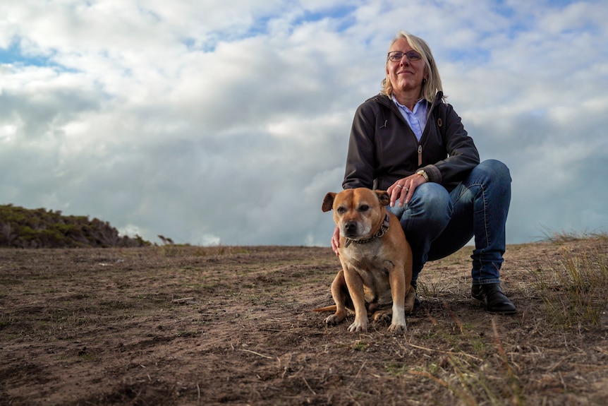 A woman wearing jeans and a grey jumper kneels with her dog in a brown field of dried grass with scattered clouds above her.